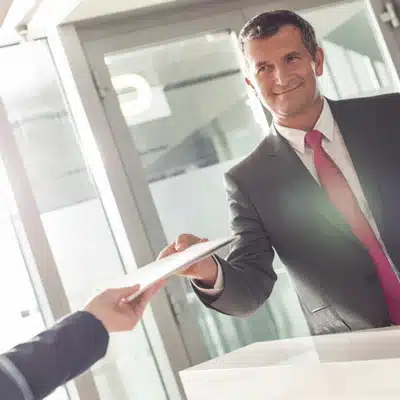 Smiling businessman in suit handing a document to another person across a reception desk