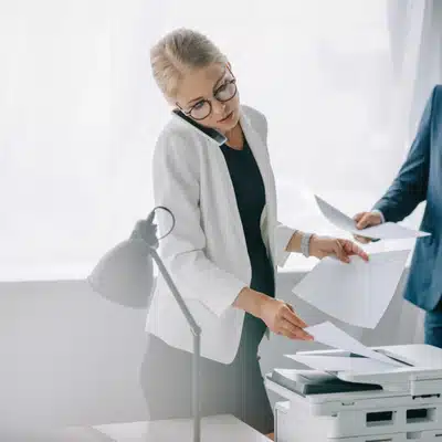 Professional on phone manages copier and documents, troubleshooting printing quality issues during urgent office tasks