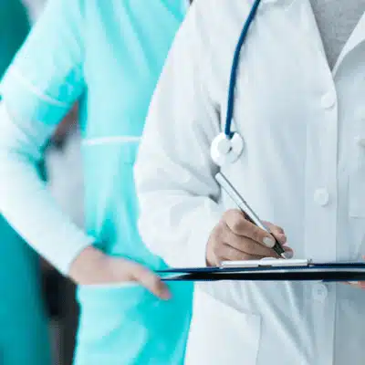 Doctor with stethoscope writes notes on clipboard, compiling medical chronology with nurse standing behind nearby