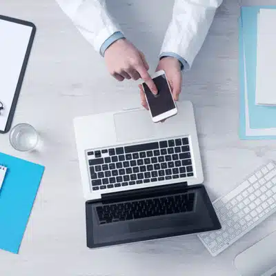 Healthcare professional uses smartphone above laptop and tablet, organizing patient records and scheduling electronic communications