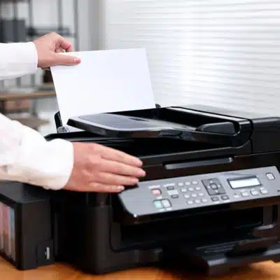 Person loads paper into multifunction inkjet printer, preparing to copy documents on wooden desk today.